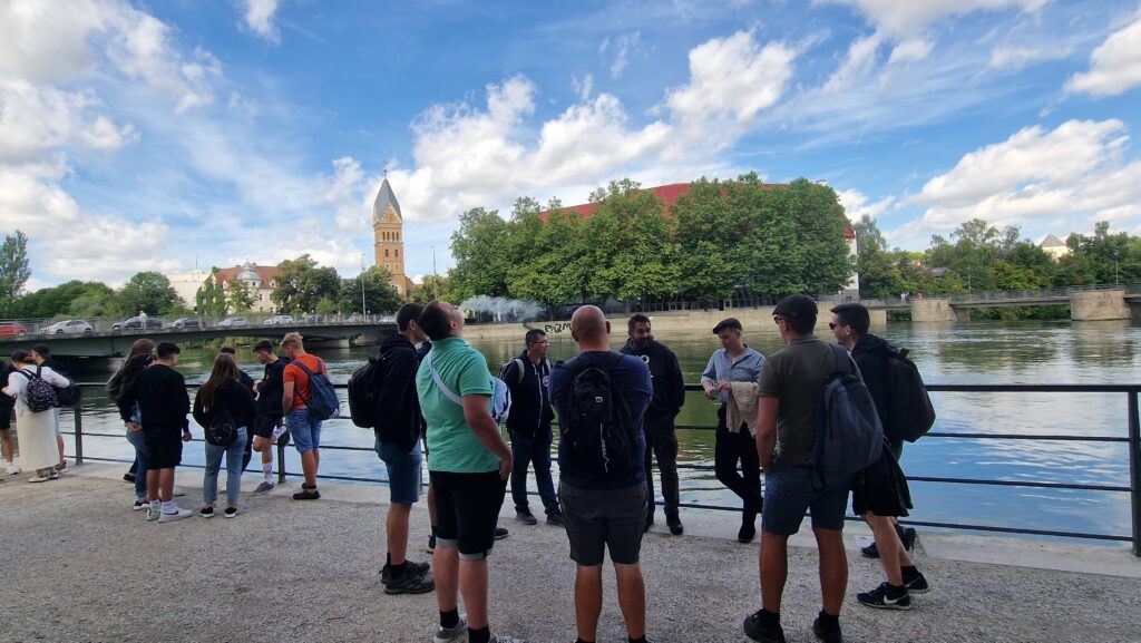Gruppe von Menschen am Flussufer, mit Kirche und Brücke im Hintergrund.