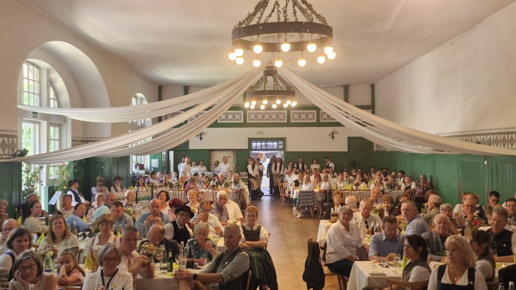 Ein festlicher Rahmen mit Gästen um Tische, dekorative Lampen und weiße Bänder.