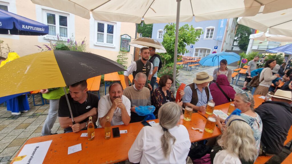Leicht bewölkt am Marktplatz in Altomünster. Menschen unter Sonnenschirmen und Tischlern im Freien.