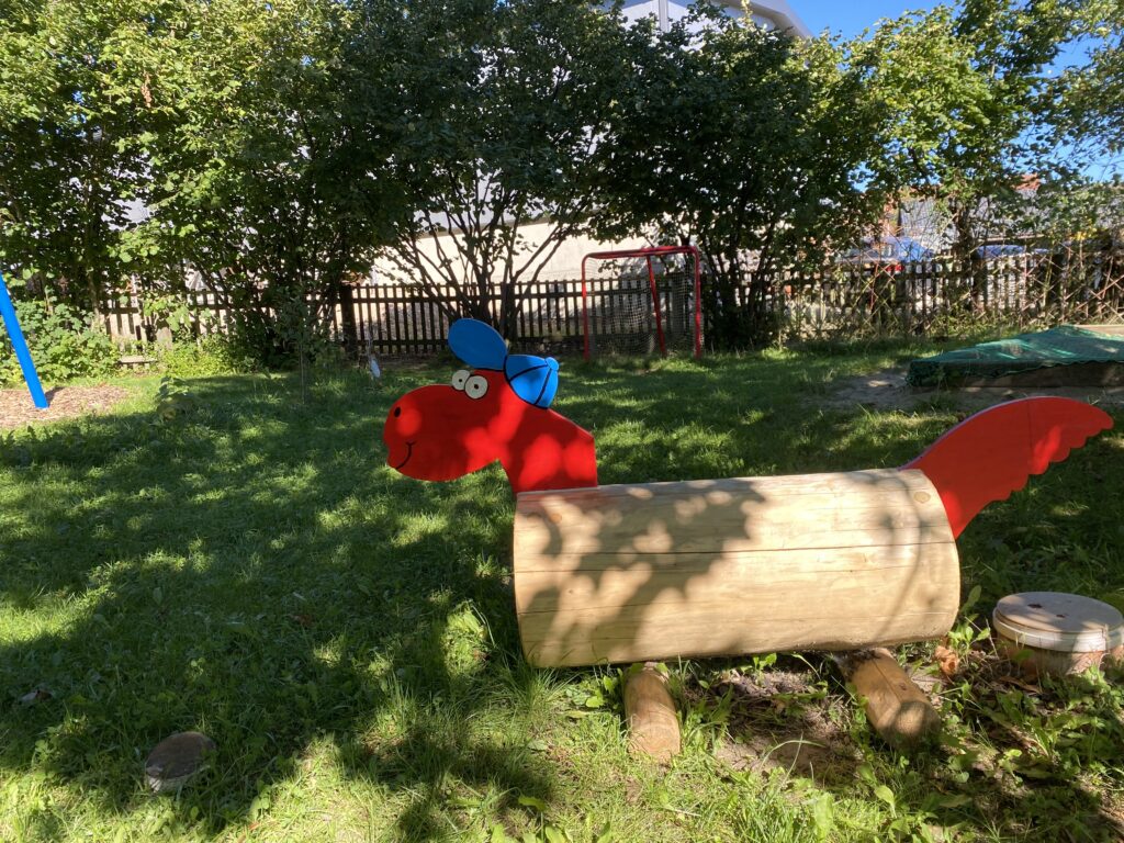 Rote Holzfigur mit Flügel und blauem Kappen, Spielplatz im Garten.