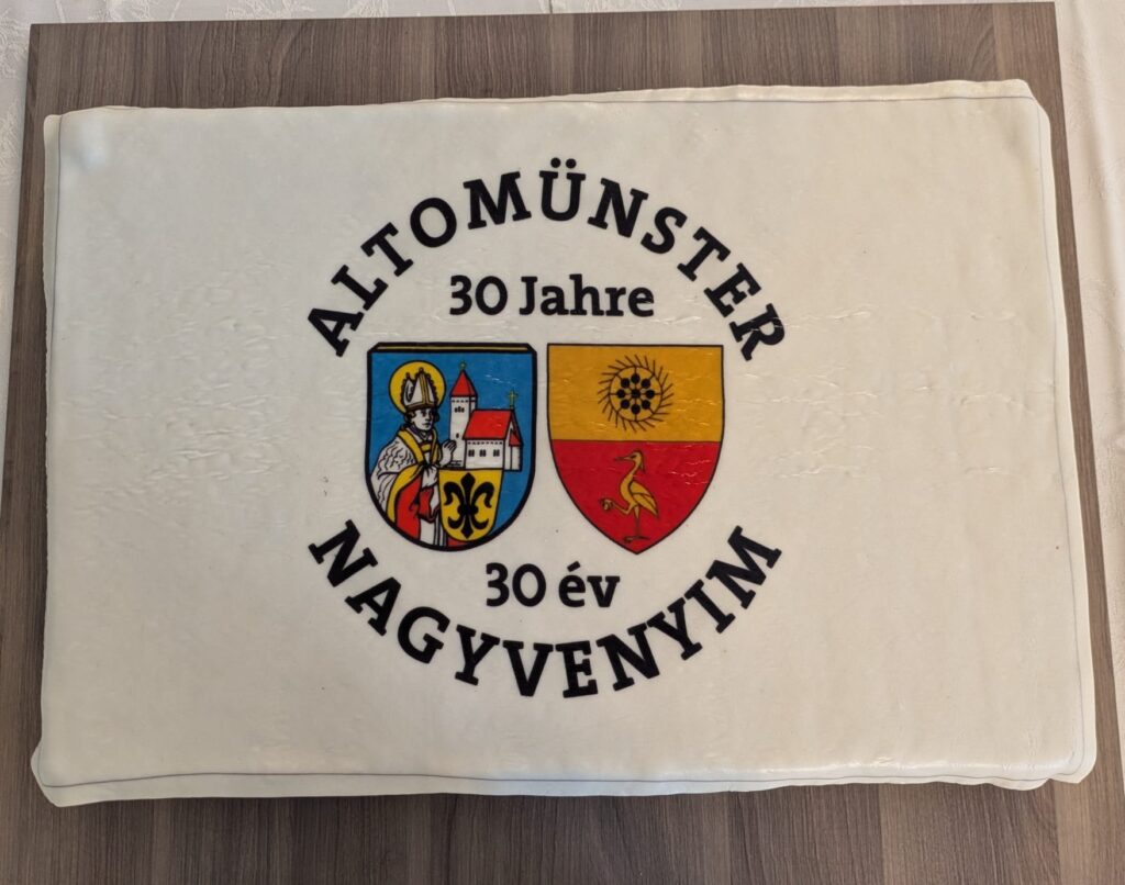 Einfaches Bild eines Kuchens mit Aufdruck „Altomünster 30 Jahre, 30 év Nagyvenyim“ auf einem Holztisch.
